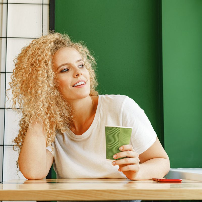 Curly young woman sitting at the table in coffee shop and relaxing with cup of coffee, portrait